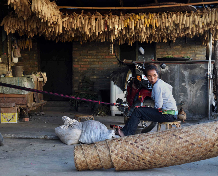 A local woman weaves a cloth shoulder bag in the entrance of her home under rows of drying maize supported by bamboo poles.  Colorful, home woven cloth shoulder bags decorated with bright fringe, tassels, or pompoms are popular cultural markers among both men and woman for ethnic minority groups like the Wa and Lahu. (Photo: Naomi Hellmann)