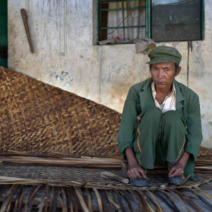An elderly man squats in the entrance of his home, weaving a stiff, flat, straw mat for drying grain on. (Photo: Naomi Hellmann)