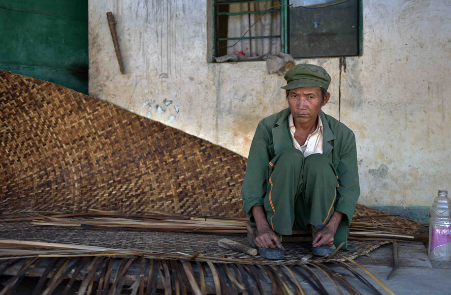 An elderly man squats in the entrance of his home, weaving a stiff, flat, straw mat for drying grain on. (Photo: Naomi Hellmann)