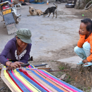 A young girl socializes with a village woman hand weaving a colorful blanket in the open space outside her home. Women often weave outside in late autumn when the air is still warm and dry and the harvest is almost over. (Photo: Naomi Hellmann)