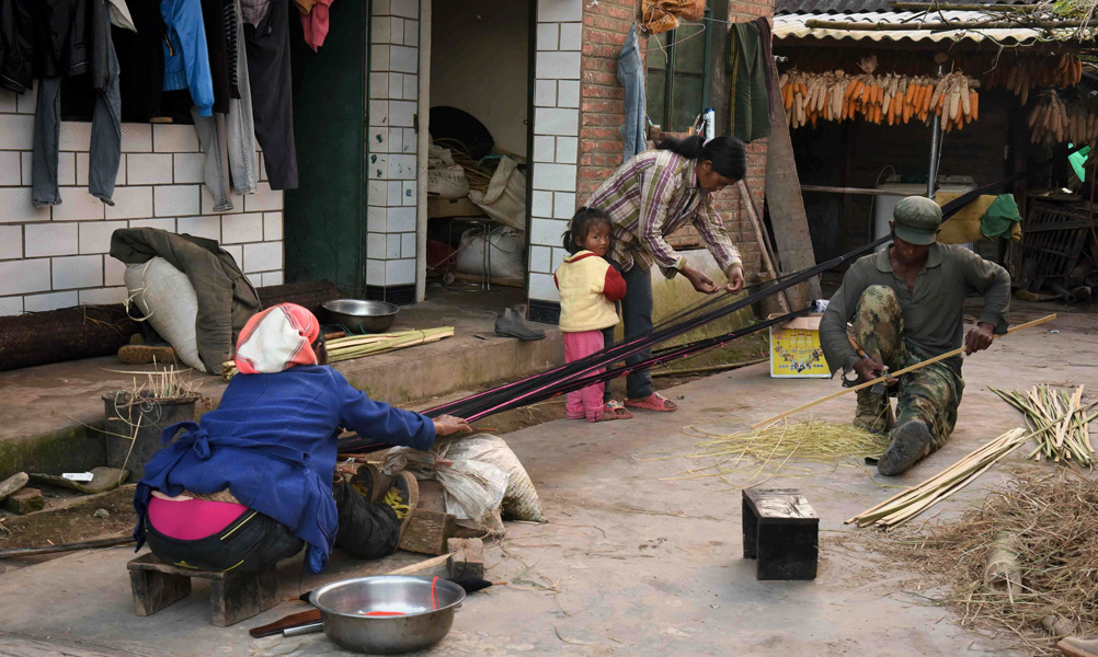 A woman and young child socialize with a neighbor weaving a cloth shoulder bag and her husband, who is splitting thin strips of bamboo with a handheld scythe, in the space outside their home. (Photo: Naomi Hellmann)