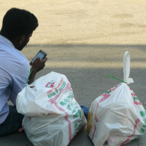 An Indian migrant, sits on the sidewalk outside the train (MRT) station texting on his mobile phone. It is a popular meeting spot, especially in the evenings and on weekends. Bags of shopping from Little India could contain gifts for family back home, or groceries for the week. (Photo: Laavanya Kathiravelu)