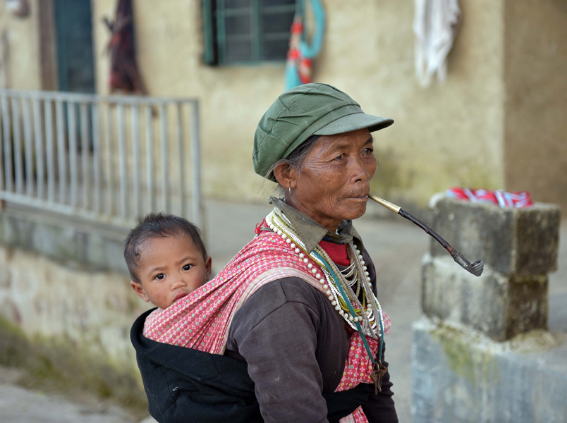 A village woman, smoking tobacco from a traditional long pipe and wearing amulets around her neck, carries a young child on her back. Children typically remain in the care of elderly extended family while their parents migrate out to distant coastal provinces for work. (Photo: Naomi Hellmann)