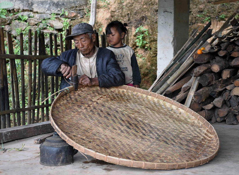 A young girl observes an elderly man fasten the rim of an open, circular, shallow, handwoven bamboo receptacle used for drying crops such as grain, tea, tobacco, fruits, spices, nuts, beans, and seeds on. (Photo: Naomi Hellmann)